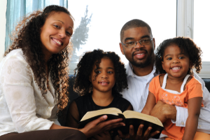 A black family, mom, dad, and two daughters sit together on a couch to watch worship online.